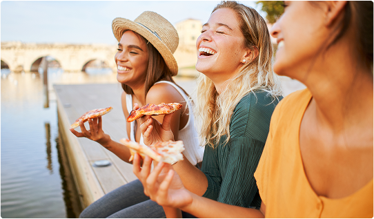 Three friends laughing and sharing pizza outdoors by the water