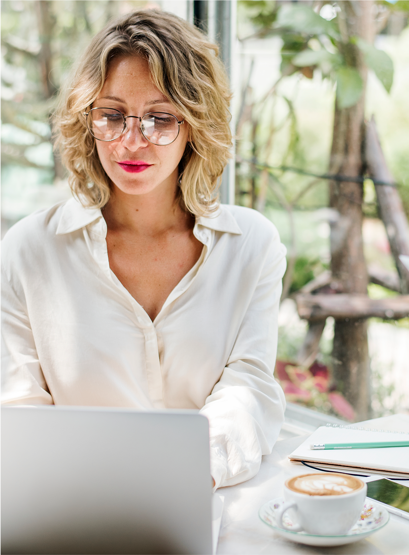 Woman with glasses working on a laptop at a sunlit table with coffee